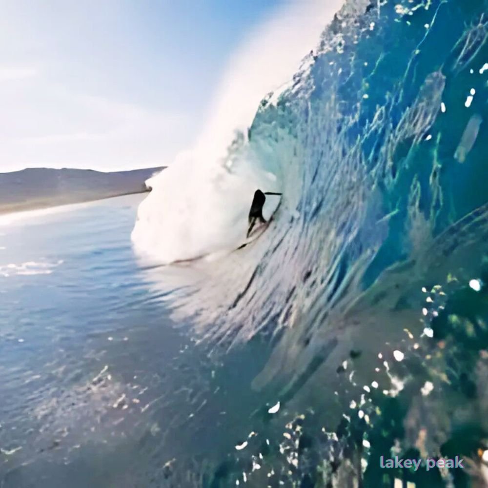 surfer is riding under a large wave that forms a water cave around them, with a beach view in the background.@lakey peaks