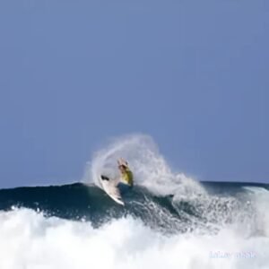 A surfer in a yellow shirt performs a maneuver on a large wave, surrounded by white ocean spray against a clear blue sky.